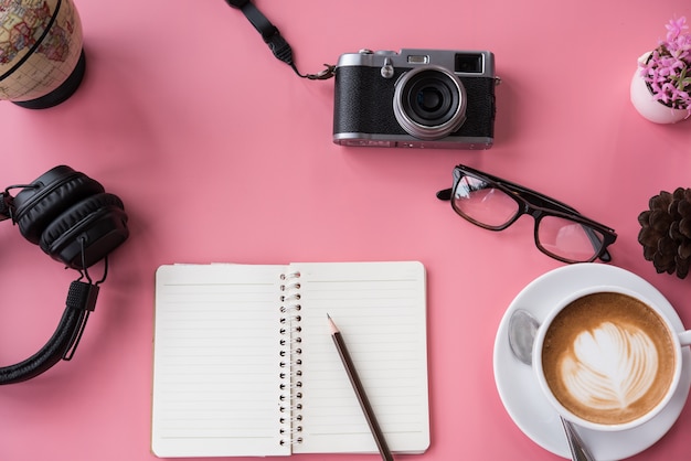 写真 カメラ、眼鏡、ヘッドフォン、メモ帳、ピンクの背景、旅行プランナーのコンセプト。
