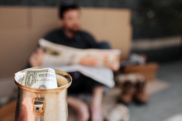 Camera is focused on metal cup with dollar in it. It is beggar's cup. Homeless man is sitting on cardboard and reading newspaper. He is relaxing.