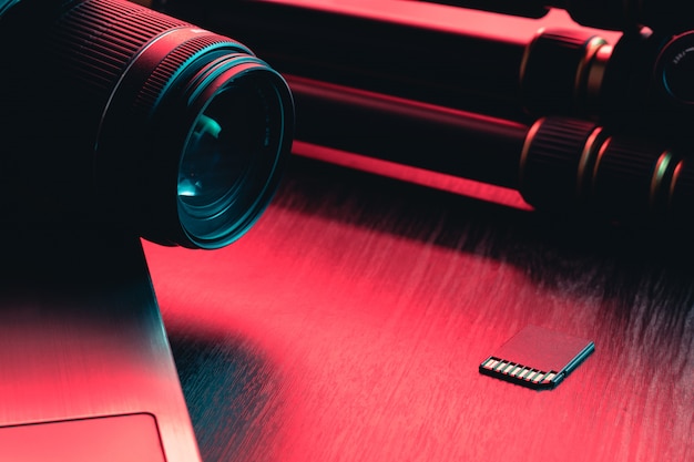 Camera lens, card, laptop and tripod on wood table. Desk workspace. Red and blue light