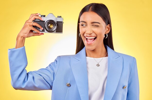 写真 スタジオでの仕事のためのカメラ写真と笑顔とウィンクを持つ女性の写真創造的な記憶と写真家のスキルを持つ面白い絵文字の顔を持つ幸せな若い女性と黄色の背景