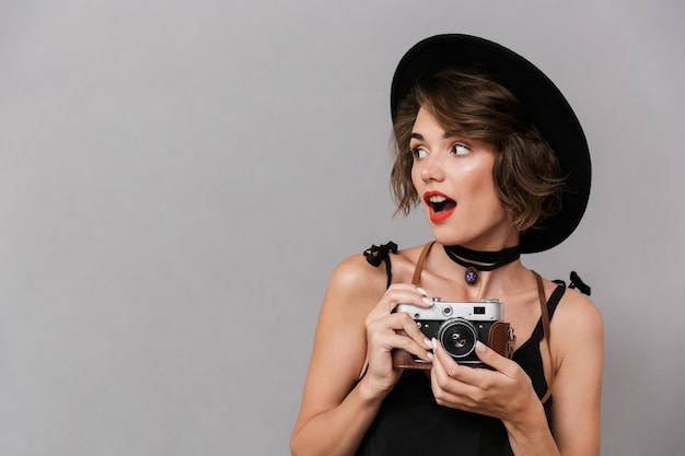 camerawoman wearing black dress and hat holding retro camera, isolated over gray wall