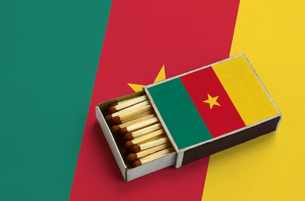 Флаг Камеруна отображается в открытой спичечной коробке, которая заполнена спичками и лежит на большом флаге