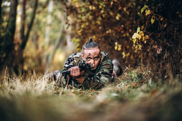 camouflage soldier with rifle and painted face lying in the grass aiming at the rifle.