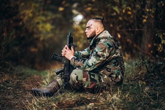 camouflage soldier with rifle and painted face sitting in the grass.