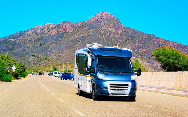 Camper or minivan on road at Costa Smeralda in Sardinia Island, Italy summer. Caravan motorhome on holidays at highway. Mini van rv on motorway. Olbia province. Mixed media.