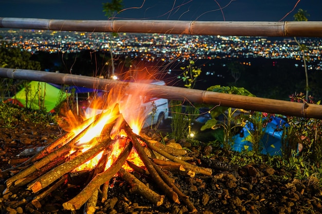 キャンプファイヤーが夜空に照らされ、街の明かりが背景に見えます。