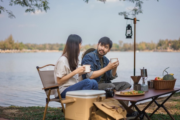 캠핑 컨셉 젊은 커플 배 여행객들이 호수 근처에 캠핑하는 동안 대화하고 커피를 마시고 있습니다.