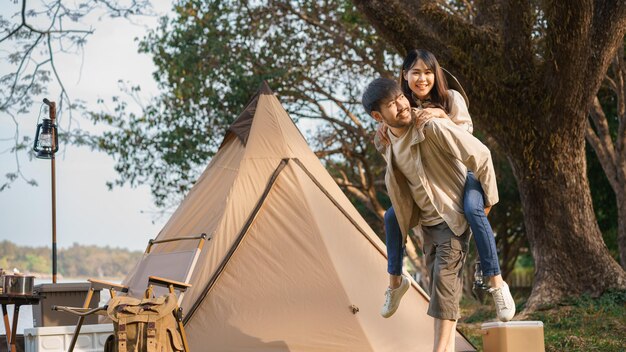 キャンプのコンセプト 若いカップルがおんぶしてガール フレンドを運び、湖の近くを歩いて景色を見ます