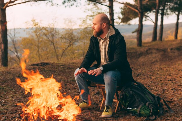 Camping, hiking, lifestyle. Hipster hiker sitting near bonfire in forest