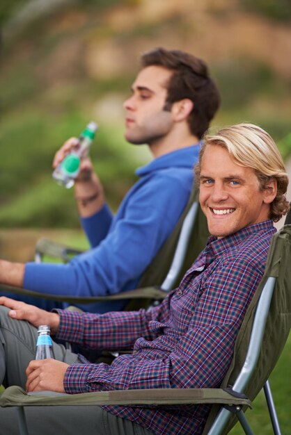 Photo camping man and portrait with drinks for relax on grass in nature with happiness for holiday and vacation people smile and alcohol at campsite in woods for adventure travel and bonding in forest