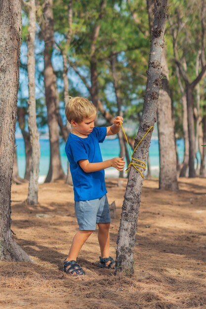 Camping mensen buiten levensstijl toeristen in zomer bos in de buurt van lazur zee Blonde serieuze jongen in blauwe tshirt studie overlevingstechnieken praktijk methoden voor het binden van touw knopen Natuurlijke kinderen onderwijs