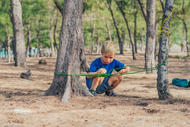 Camping mensen buiten levensstijl toeristen in zomer bos in de buurt van lazur zee Blonde serieuze jongen in blauwe tshirt studie overlevingstechnieken praktijk methoden voor het binden van touw knopen Natuurlijke kinderen onderwijs