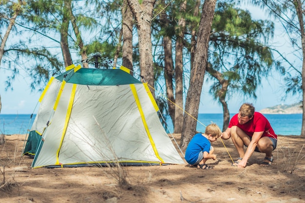 Camping people outdoor lifestyle tourists put up set up green grey campsite summer forest near lazur sea Boy son helps father study mechanism of modern easy to fold tent Natural children education