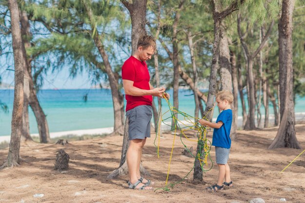 Camping people outdoor lifestyle tourists in summer forest near lazur sea Blond boy son with father study survival techniques practice methods of tying rope knots Natural children education