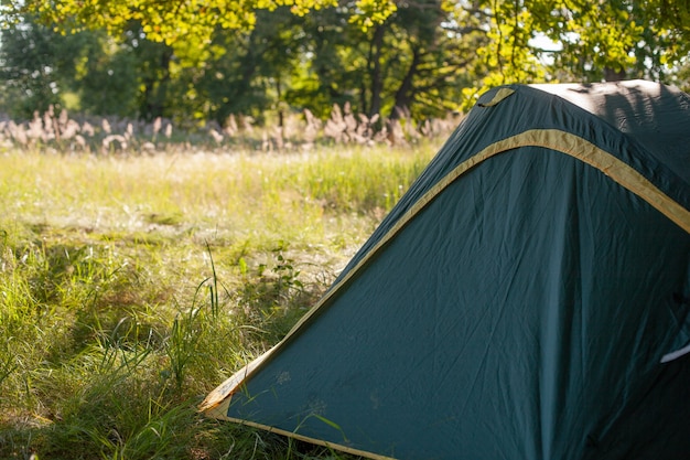 夏の輝く朝の日の出の間に森の近くのオープンフィールドの真ん中にキャンプテント。アウトドアキャンプアドベンチャーのコンセプト