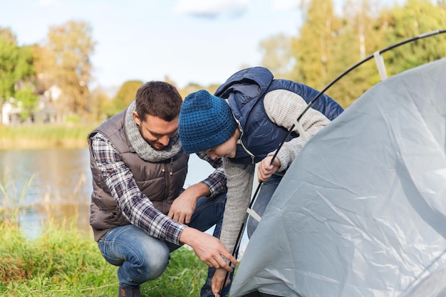 캠핑, 관광, 하이킹, 가족 및 사람 개념 - 행복한 아버지와 아들이 야외에서 텐트를 치고 있습니다.