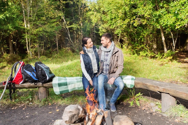 camping, travel, tourism, hike and people concept - happy couple sitting on bench and hugging near campfire at camp in woods