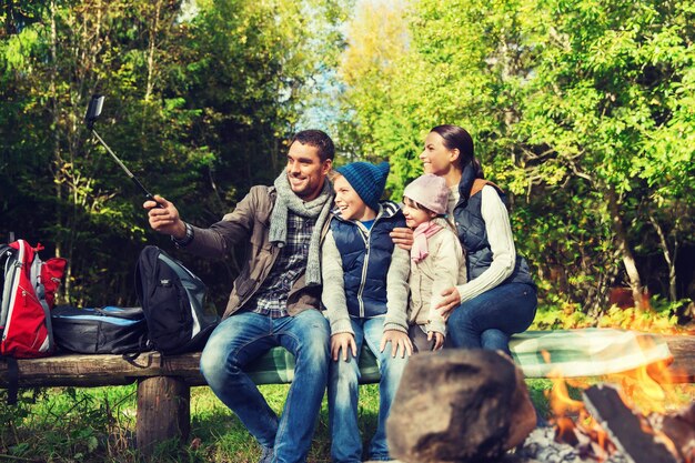 Photo camping, travel, tourism, hike and people concept - happy family sitting on bench and taking picture with smartphone on selfie stick at campfire in woods