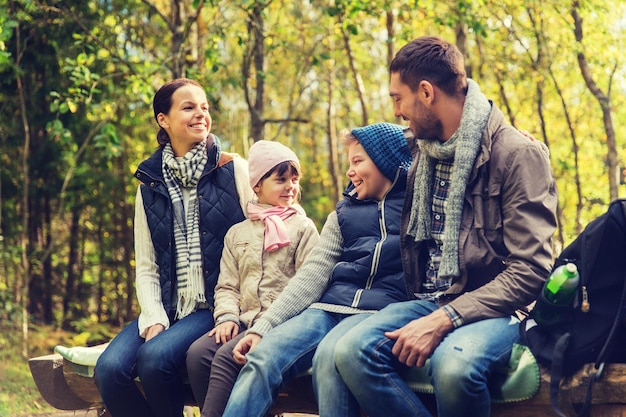 camping, travel, tourism, hike and people concept - happy family sitting on bench and talking at camp in woods