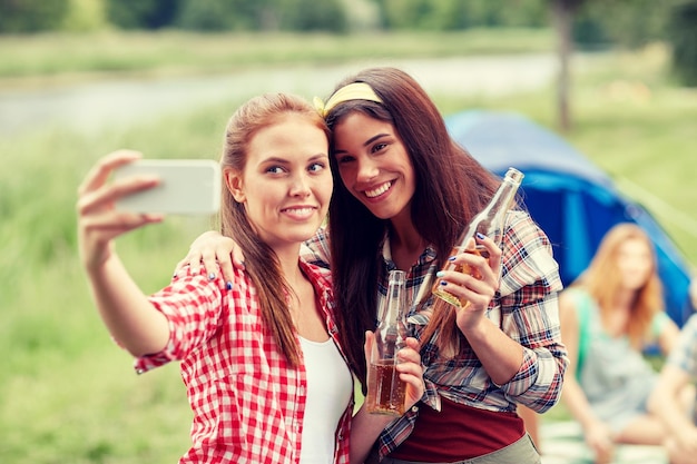 camping, travel, tourism, hike and people concept - happy young women with glass bottles drinking cider or beer and taking selfie by smartphone at camping