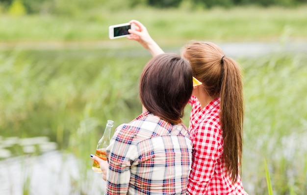 camping, travel, tourism, hike and people concept - happy young women with glass bottles drinking cider or beer and taking selfie by smartphone outdoors from back