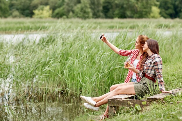 camping, travel, tourism, hike and people concept - happy young women with glass bottles drinking cider or beer and taking selfie by smartphone outdoors