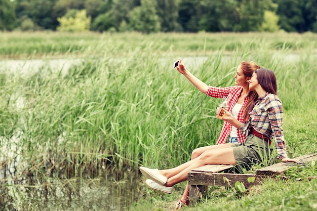 camping, travel, tourism, hike and people concept - happy young women with glass bottles drinking cider or beer and taking selfie by smartphone outdoors