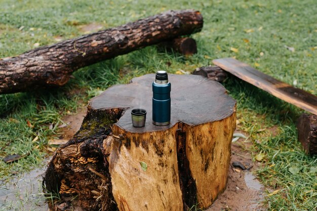 Camping vacuum thermos and mug of hot drink standing on stump table at camping site in rainy autumn weather. Hiking, camping, warming drink in cold season concept.