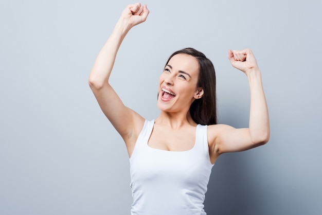 Can not hide her emotions. Beautiful young women keeping arms raised and smiling while standing against grey background