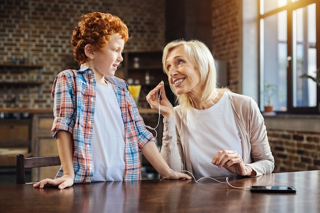 Photo can i try. positive minded senior woman smiling to her little grandson while both spending time together and listening to music at home.