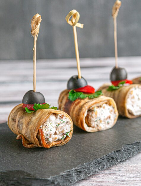 Canapes of fried eggplant stuffed with cheese, herbs. Garnished with sweet pepper, parsley and olive. On a black slate board. Gray background. Close-up.