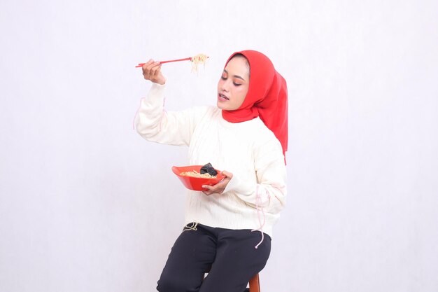 candid asian woman wearing hijab looking down holding noodles with chopsticks from the side and bowl