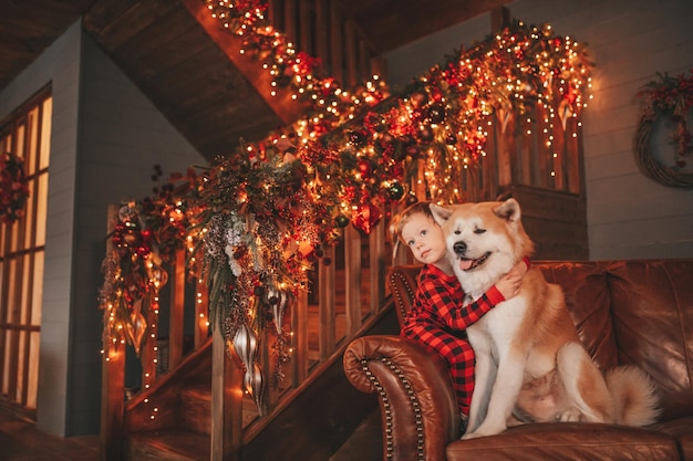 赤い格子縞のパジャマを着た率直な本物の幸せな少年は、クリスマスの装飾が施されたロッジで蝶ネクタイをした犬を抱きしめる