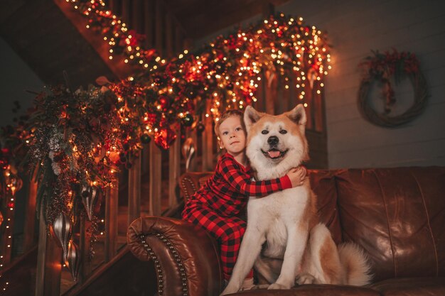 Candid authentic happy little boy in red plaid pajama hugs dog with bow tie at lodge Xmas decorated