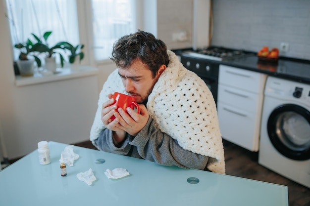 Candid beeld van een gestresste jonge man in een deken die thuis met griep te maken heeft en op een keuken zit