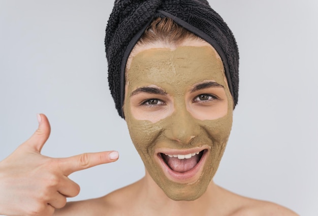 Candid closeup portrait of happy young woman indicate with finger on her cosmetic green clay organic mask on her face wears black towel on hair Female take care of face skin isolated on white wall