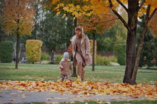 Foto la famiglia candida corre attraverso le foglie gialle autunnali. madre e bambino che ridono e si divertono nel parco naturale. modelli che indossano maglione beige, giacca, berretto