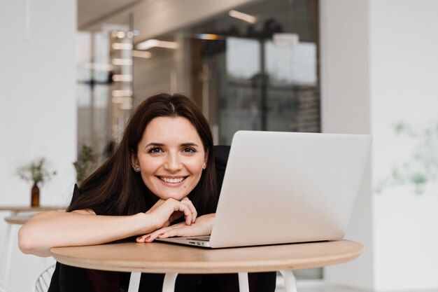 Candid girl with laptop sits in white cafe smiles and rejoices\
at successful work in it company cheerful young woman programmer\
works remotely on laptop and try to meet deadline