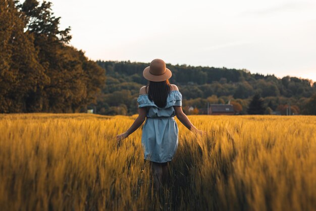 Photo candid portrait of a brunette aged 20-24 walks in blue dress and hat in cornfield smiling naturally