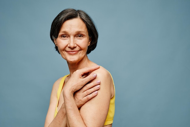 Candid portrait of mature woman smiling at camera while posing elegantly against pastel blue backgro
