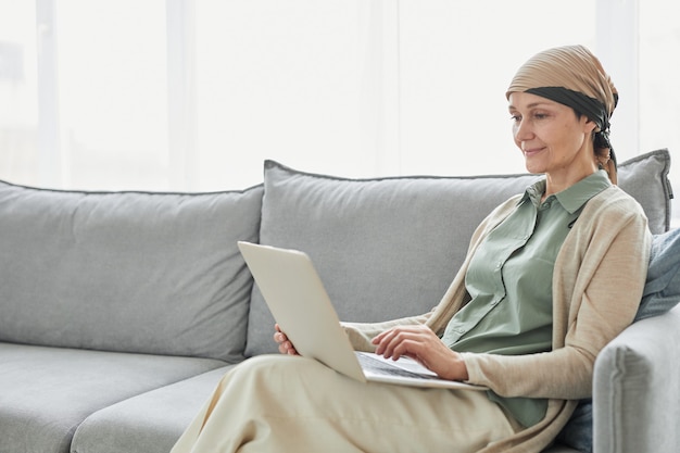 Candido ritratto di donna matura che usa il velo e usa il laptop mentre si rilassa sul divano in un accogliente ambiente domestico, spazio copia