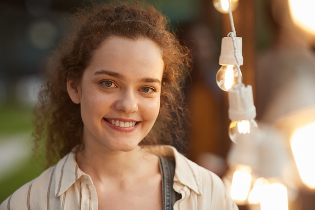 Candid portrait of smiling young woman standing by lights at outdoor party