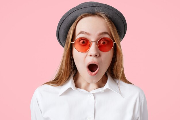 Candid shot of amazed terrified female with wide opened mouth, dressed in white shirt, black stylish hat, poses against pink wall Emotional beautiful surprised teenager stares at camera