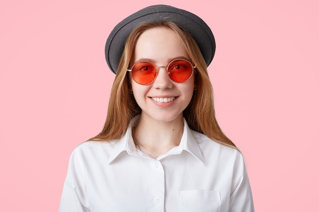 Candid shot of beautiful female with cheerful expression, wears trendy red shades and black hat, smiles joyfully, rejoices having vacation, isolated over pink wall People, style, happiness