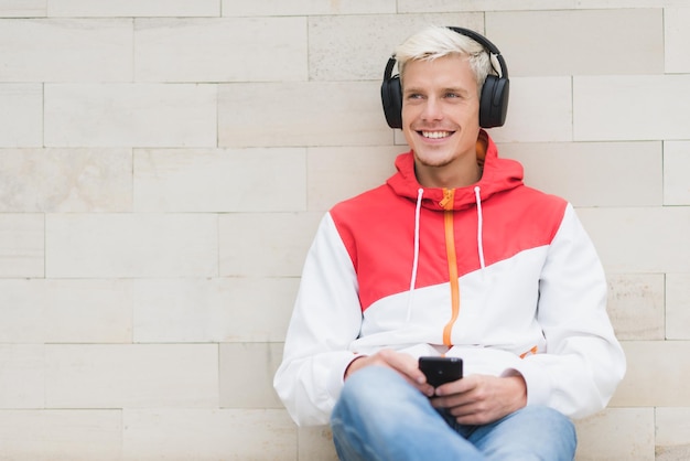 Foto colpo schietto di felice maschio sorridente vestito con abiti sportivi rossi rilassarsi e sedersi dopo attività fisiche all'aria aperta nel parco bell'uomo che ascolta la musica con le cuffie dallo smartphone all'esterno