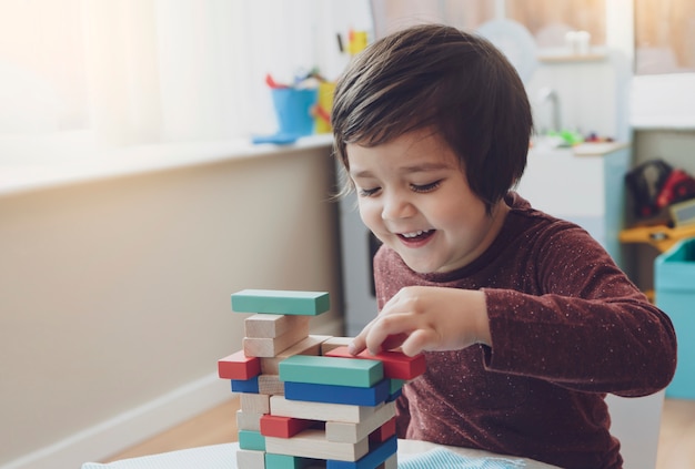 陽気な男の子の率直なショットは、プレイルームでカラフルな木製ブロックを再生し、自宅で木製のブロックを積み重ねる子供の肖像画、就学前と幼稚園の子供のための教育玩具。クリエイティブコンセプト