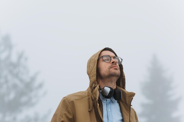 Candid shot of smart male listening to music using his headphones and mobile phone looking up and wearing hood of coat and eyeglasses during walk in the misty park People and technology concept