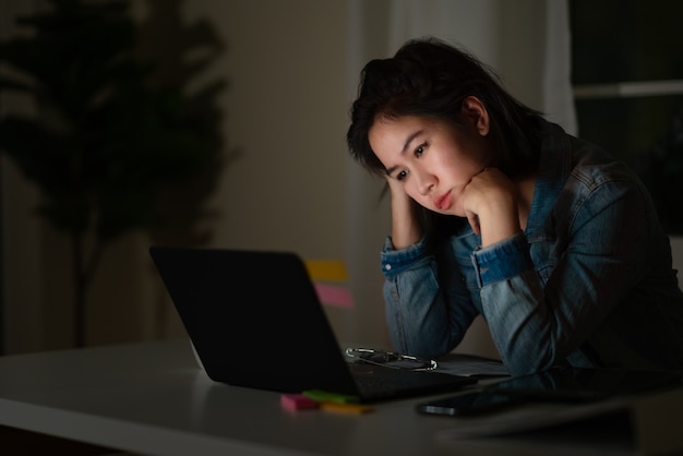 若いアジアの独身女性学生の率直な仕事は、ホームオフィスでコンピューターのラップトップまたはノートブックのプロジェクト研究問題で夜遅くまでストレスを感じます。アジア人の職業上の燃え尽き症候群の概念。