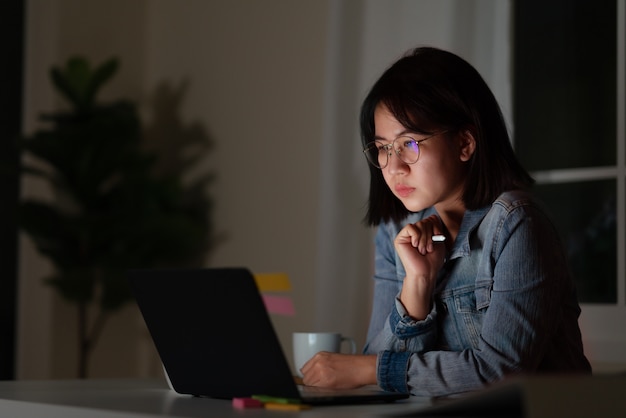 Foto candido di giovane studentessa asiatica attraente che si siede sullo scrittorio con l'aggeggio digitale astuto che esamina taccuino che funziona a tarda notte con la ricerca di progetto, il grafico o il concetto del programmatore.