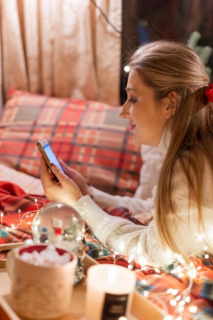 Candid young smiling woman communicates with friends using a smartphone texting message at home in a cozy bed on a red checkered blanket or plaid on happy new year and merry christmas eve